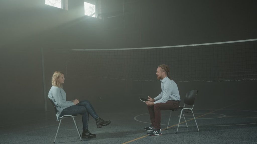A man and a woman engage in a therapy session in a smoky indoor setting, sitting apart in a large room.