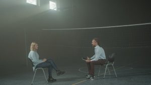 A man and a woman engage in a therapy session in a smoky indoor setting, sitting apart in a large room.